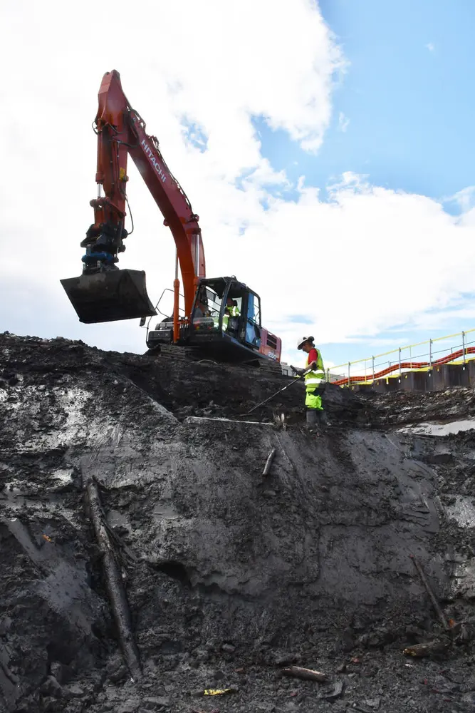 Arkeolog i gule synlighetsklær og hvit hjelm står på en avsats i en skråning av grå leire og holder en metalldetektor. En oransje gravemaskin står på kanten av skråningen.