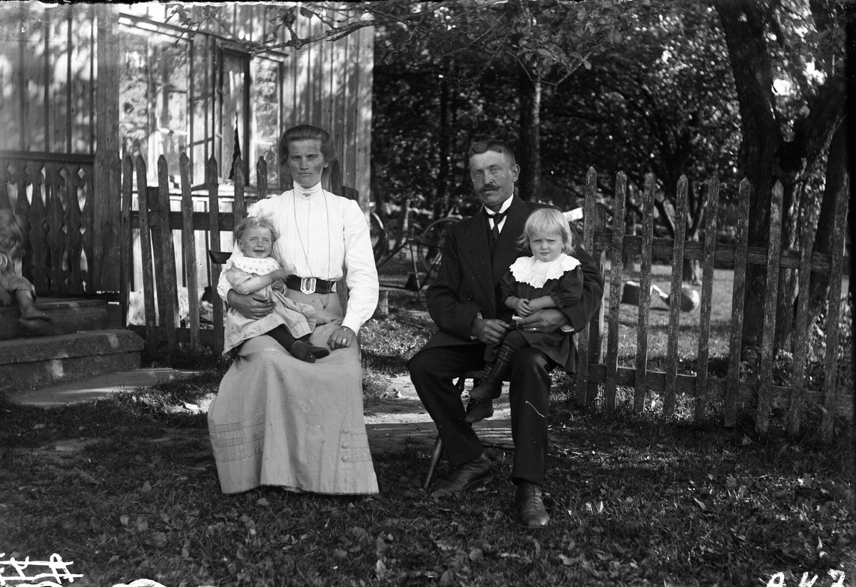 Hilma och Vilhelm Andersson sitter vid ett spjälstaket i trädgården med varsitt barn i knät och ytterligare ett skymtar till vänster på farstukvisten till huset. Barnen bär stora vita kragar. (Se även bild nr GEA076)