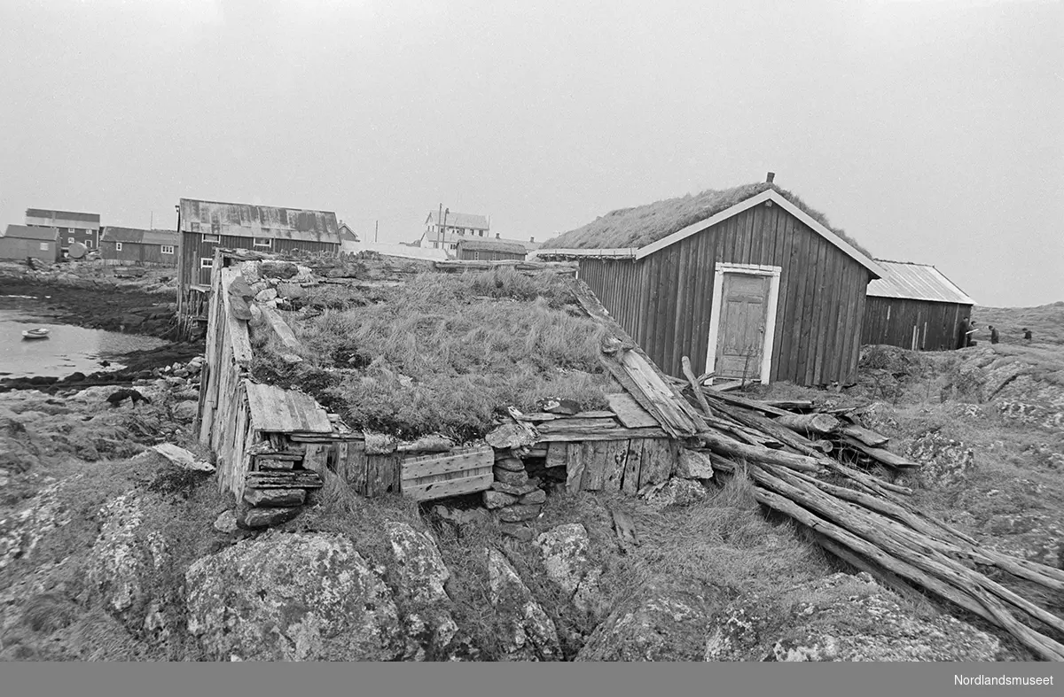 Gårdsøya med bebyggelse. Brygge, uthus. En robåt på vannet til venstre. Fire personer ved et skur til høyre i bildet. Helt i bakgrunnen boliger.