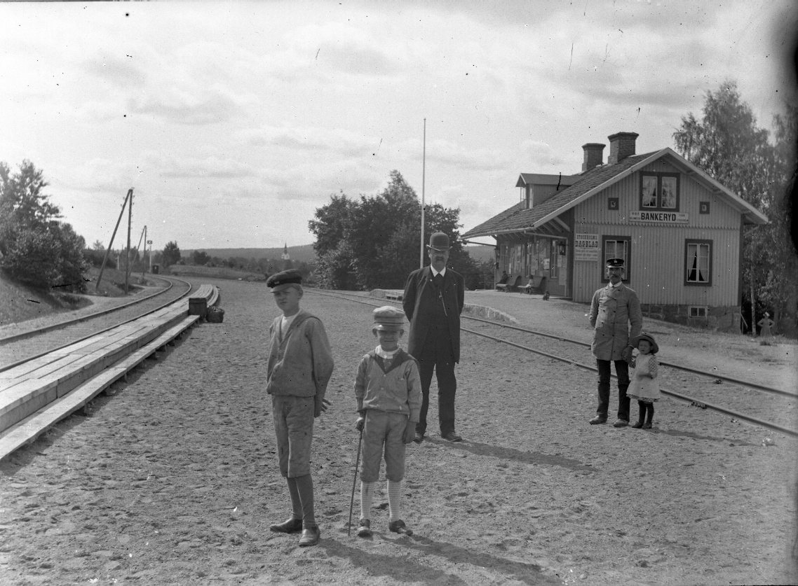 Två pojkar i sjömanskostym står framför en man med kubb och käpp framför Bankeryds järnvägsstation. Något bakom dem till höger står en annan man i uniform med en liten flicka i handen. Det är troligen stationsmästare Gustaf Hessel.