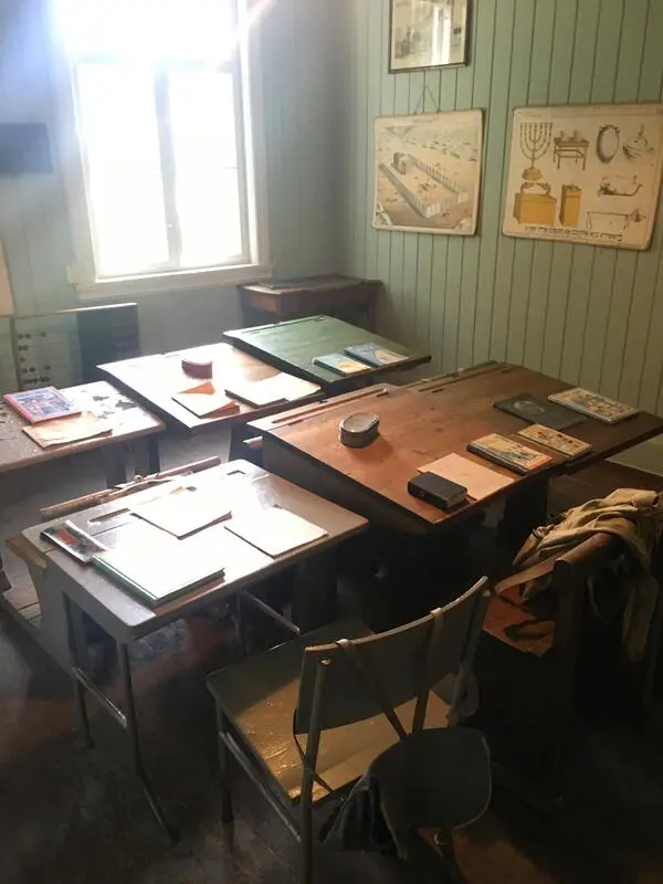 Skinnarbøl skolemuseum. Fotografi tatt inne i klasserommet. Veggen er lysegrønn, på den henger skoleplansjer. Fire gamle skolepulter.