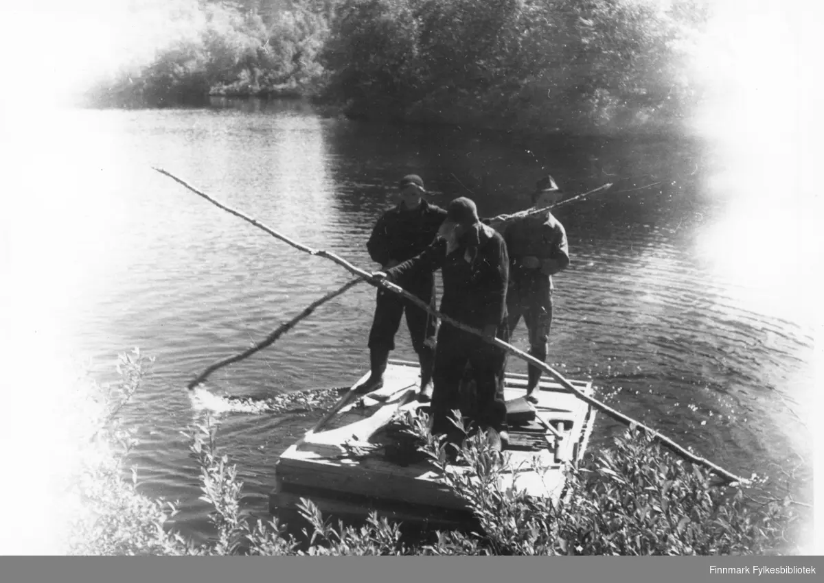 Tre menn står på en flåte ute på en elv. To av mennene har staker i hendene.