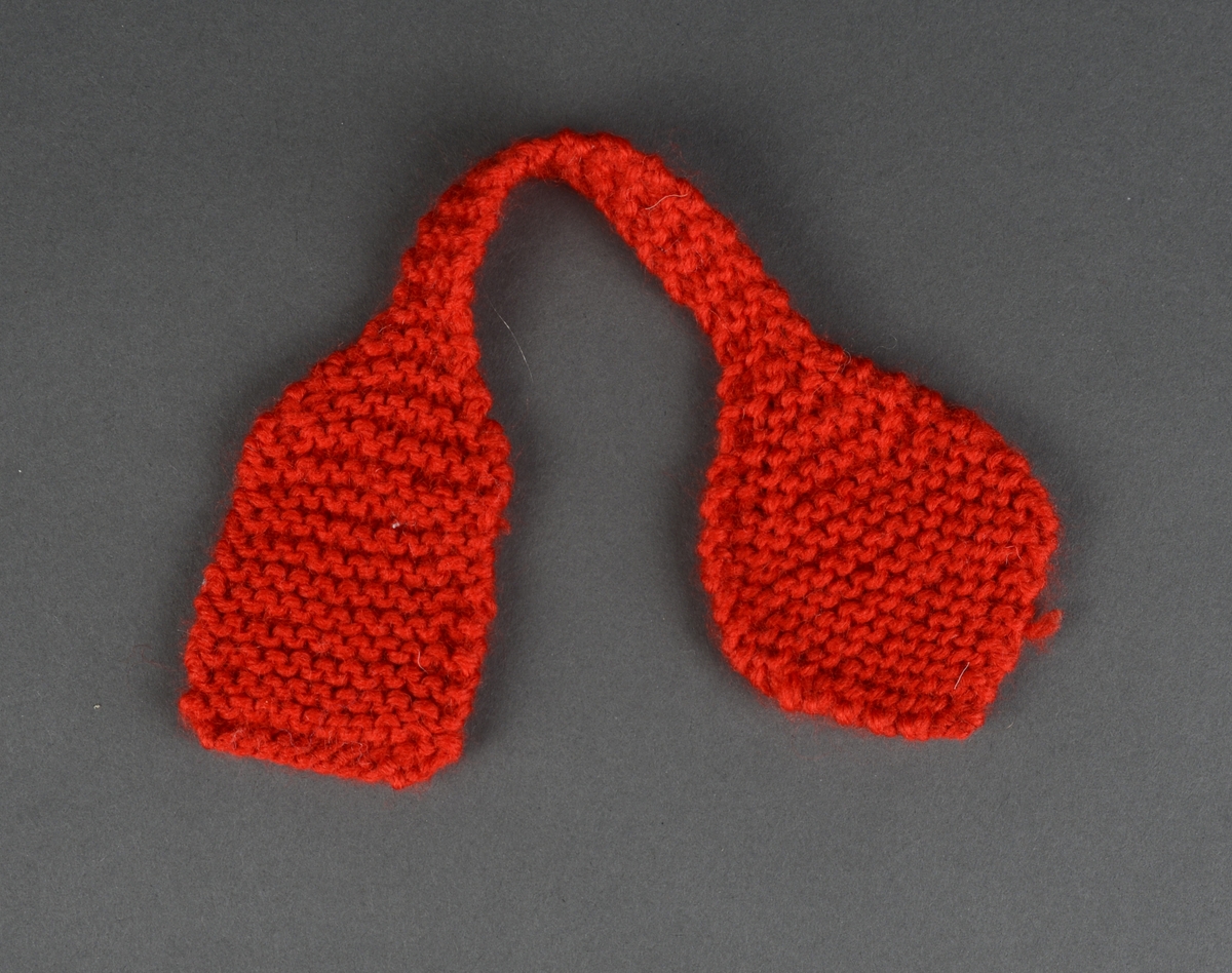Et dukkeskjerf strikket av rødt ullgarn. Midtpartiet er smalt. Den ene enden er firkantet på form, mens den andre enden er dråpeformet. Skjerfet er strikket i rillestrikk.
