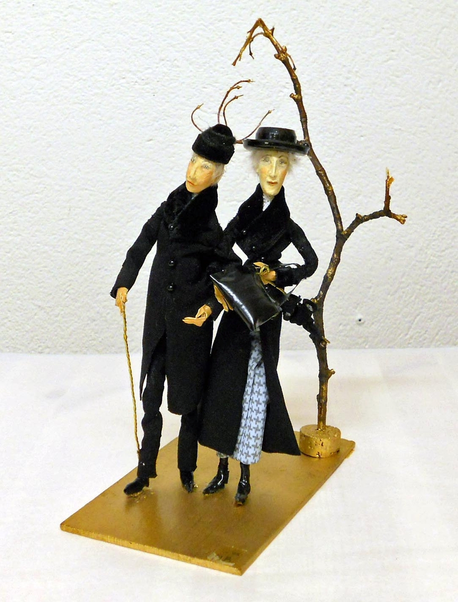Ett äldre par, klädda i svart, står arm i arm vid ett kalt träd. Han har käpp i handen, hon bär en handväska.