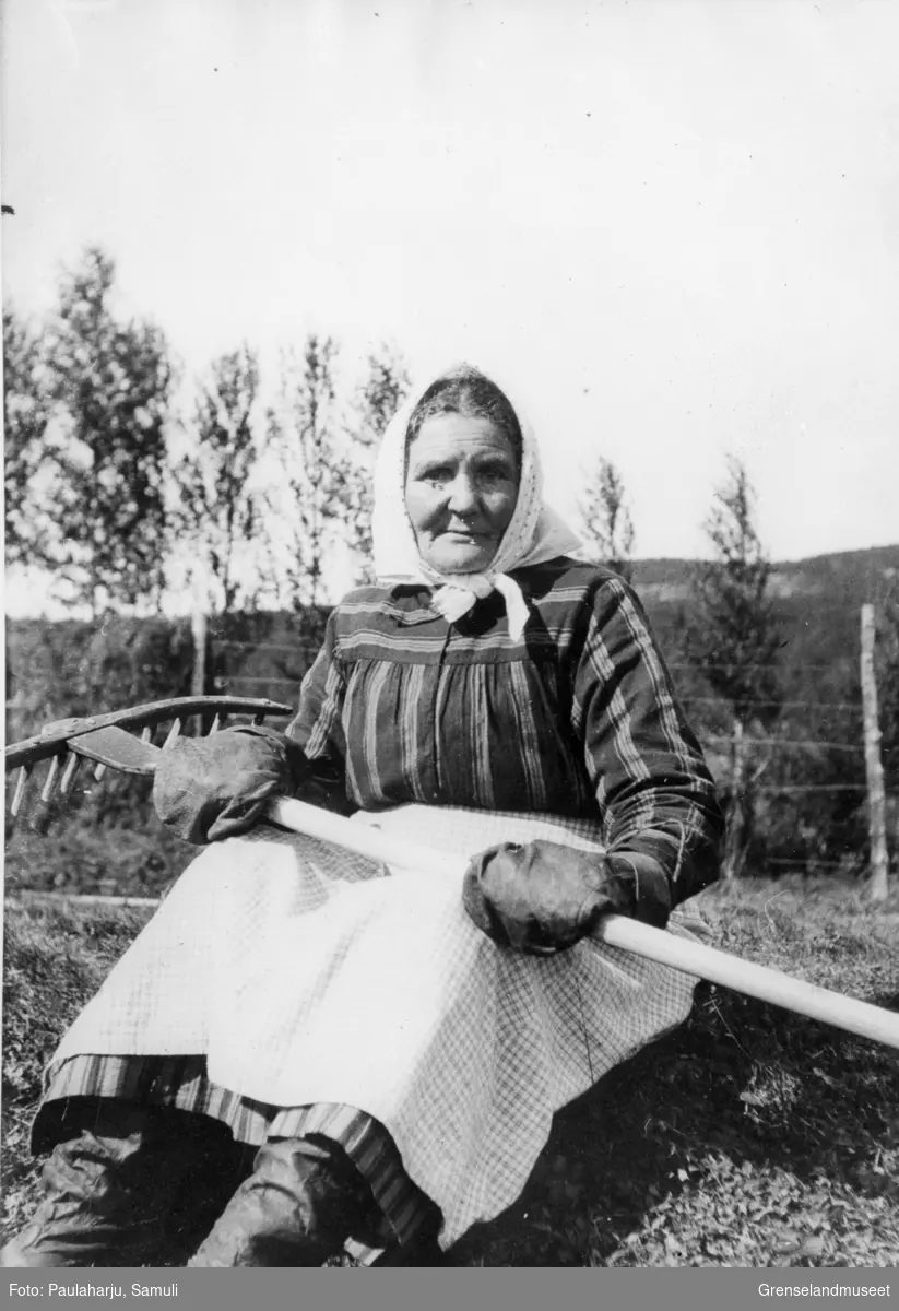 "Mikkolas hustru". En eldre kvinne sitter med en rake i hendene foran en hesje. 