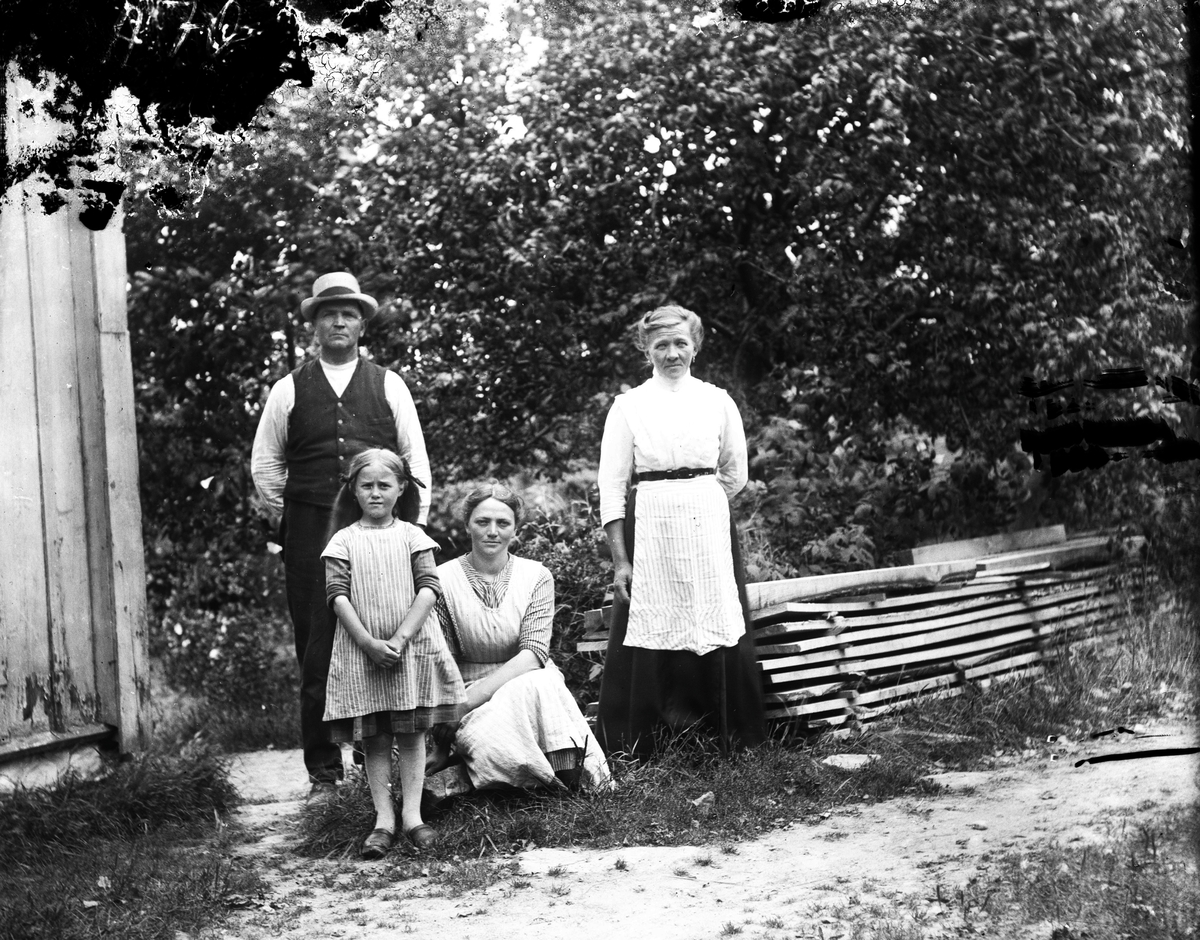 En familj med två döttrar samlad vid husknuten. Den äldre som sitter på huk är Hilda i Hornagård i Sällstorp. Till höger ligger brädor på tork.