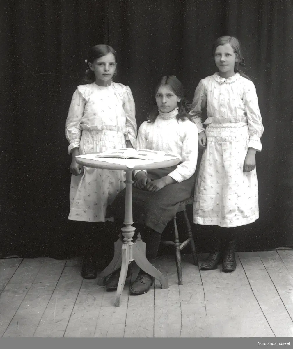 Gruppebilde, 3 jenter, den ene sitter på en stol/krakk ved et rundt bord med en bok/album oppslått, og de to andre står på hver side av henne.