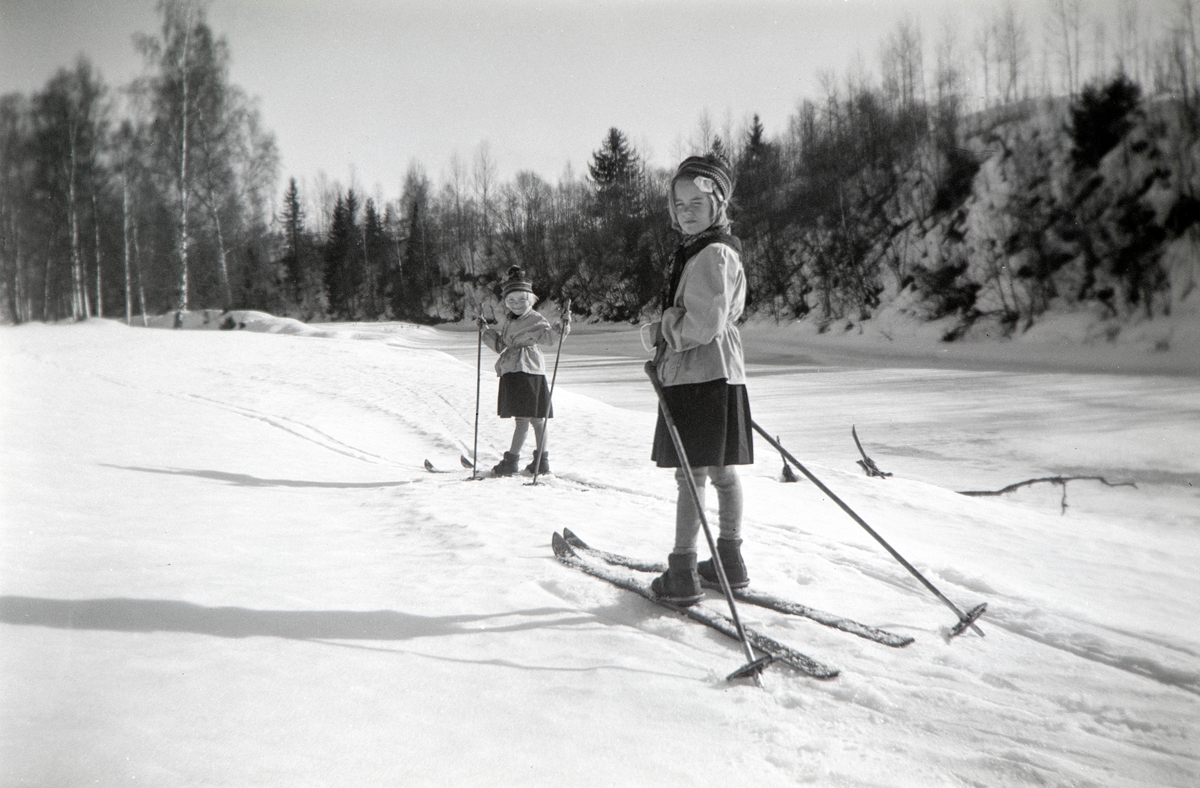 To jenter på ski fra Hol vestre i Vang til Nordre Arneberg i Romedal slutten av mars. Ikledd skjørt i anledning at de er på tur til en bursdag. Is på Svartelva. Jentene er Ragnhild Glærum (f: 1945) og Eli Vesaas (f: Glærum 1947). Fotograf Andrea Glærum (f: Gjestvang 1914)