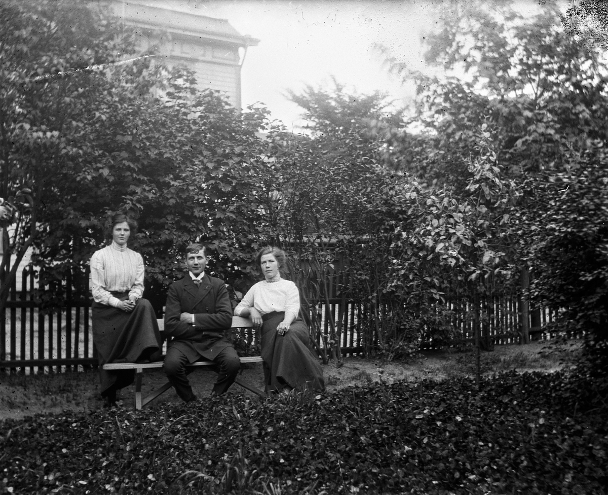 Tre ungdomar på en parkbänk vid ett spjälstaket och bakom häcken syns en stor byggnad. I mitten sitter en yngling med en kvinna på var sida.