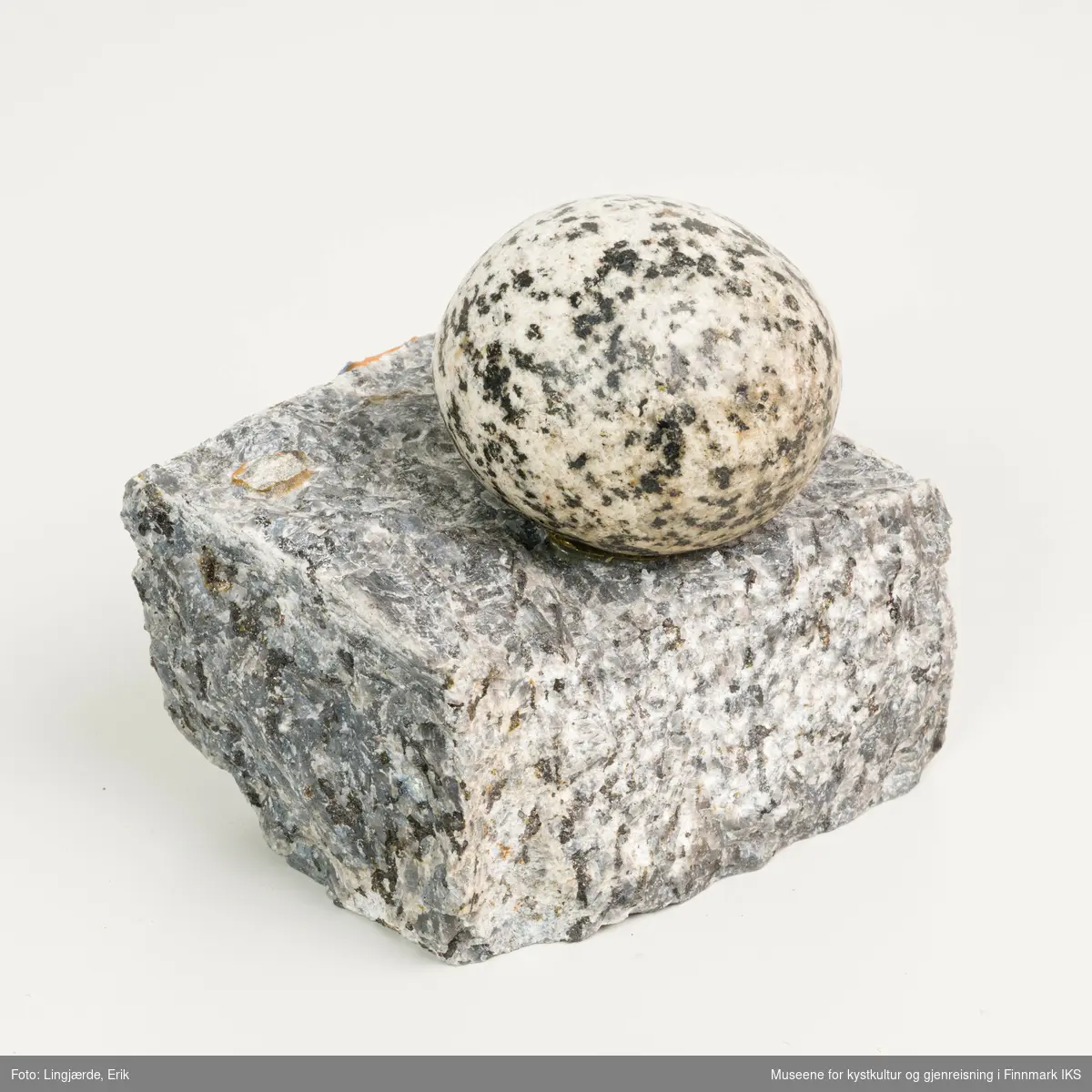En liten oval, nesten rund stein hviler på en firkantet stein, som er glatt polert på to sider og grov på de andre to sider.