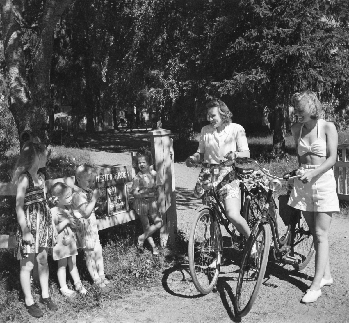 Två kvinnliga cyklister i samtal med fyra barn vid grinden till ett vandrarhem tillhörande STF.