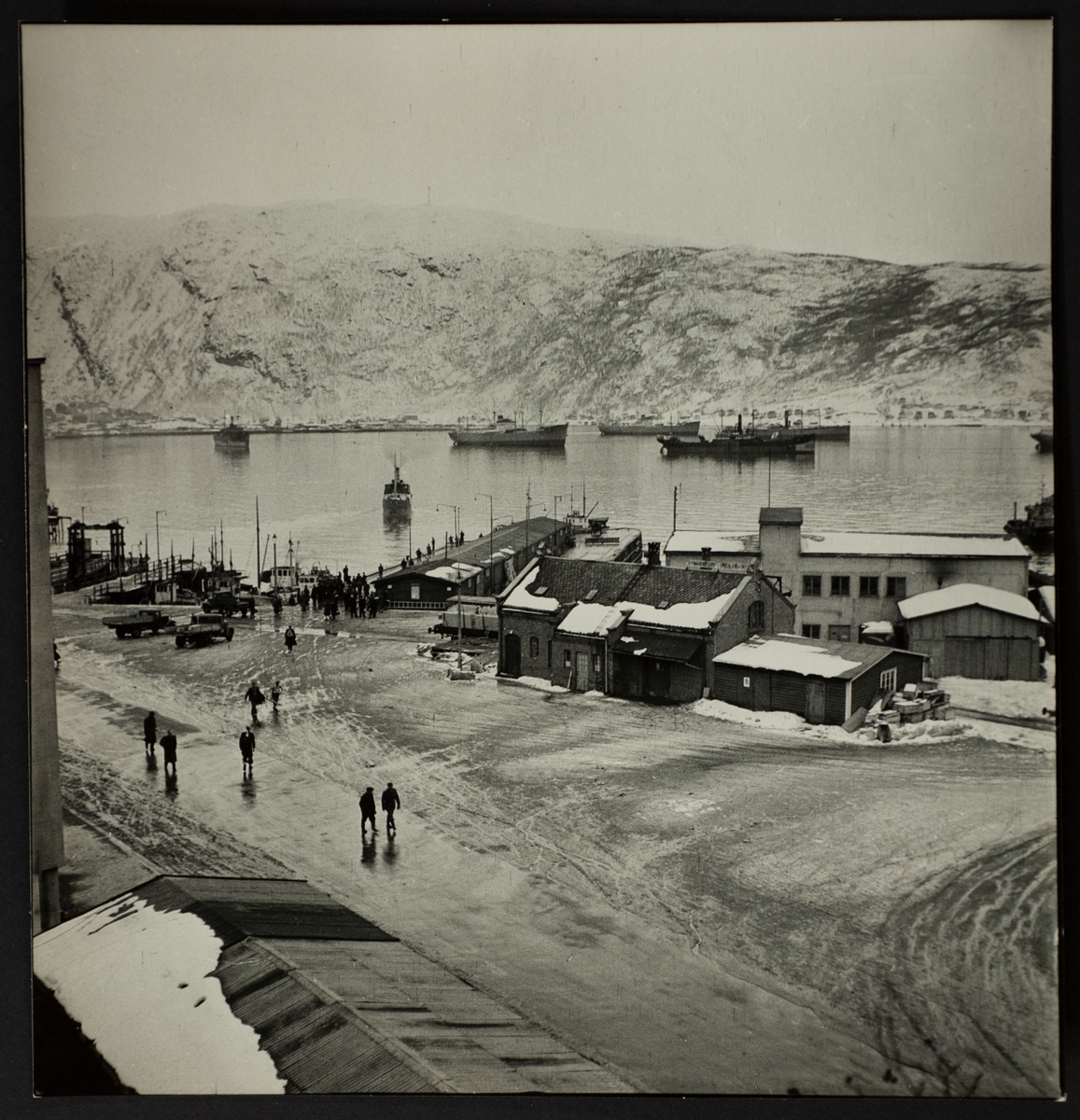 Vy över hamnplan, Narvik. Båtar ligger på redden, parkerade bilar och människor i rörelse. I bakgrunden fjäll.