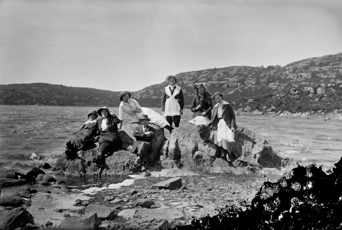 Sju personer spexar på ett par stenar i en sjö (Stamnasjön ligger i Sällstorp och i närheten även Hulta- respektive Deromesjön). Killen som står upp i mitten har dragit på sig ett förkläde ovanpå kavajen och bytt huvudbonad med kvinnan till höger. Hon har håret utsläppt, vilket inte var vanligt vid den tiden. Kvinnan som håller i hatten till vänster räcker ut tungan mot kameran och kvinnan ovanför henne intar en halvliggande pose med käppen mot hakan.
(Se även bildnr GEA147)