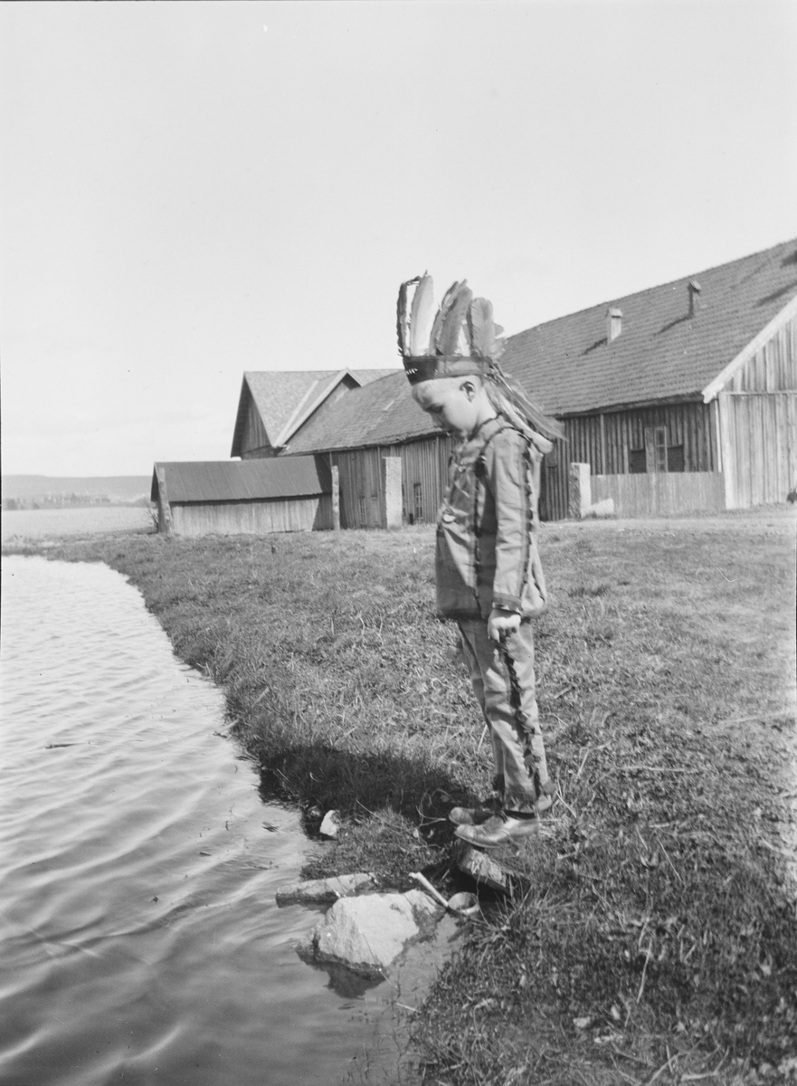 Iacob Ihlen Mathiesen står ved en gresskledd vannkant kledd i et indianerkostyme: bukse overdel og fjærpynt på hodet. I bakgrunnen sees en stor, umalt bygning med flere fløyer.