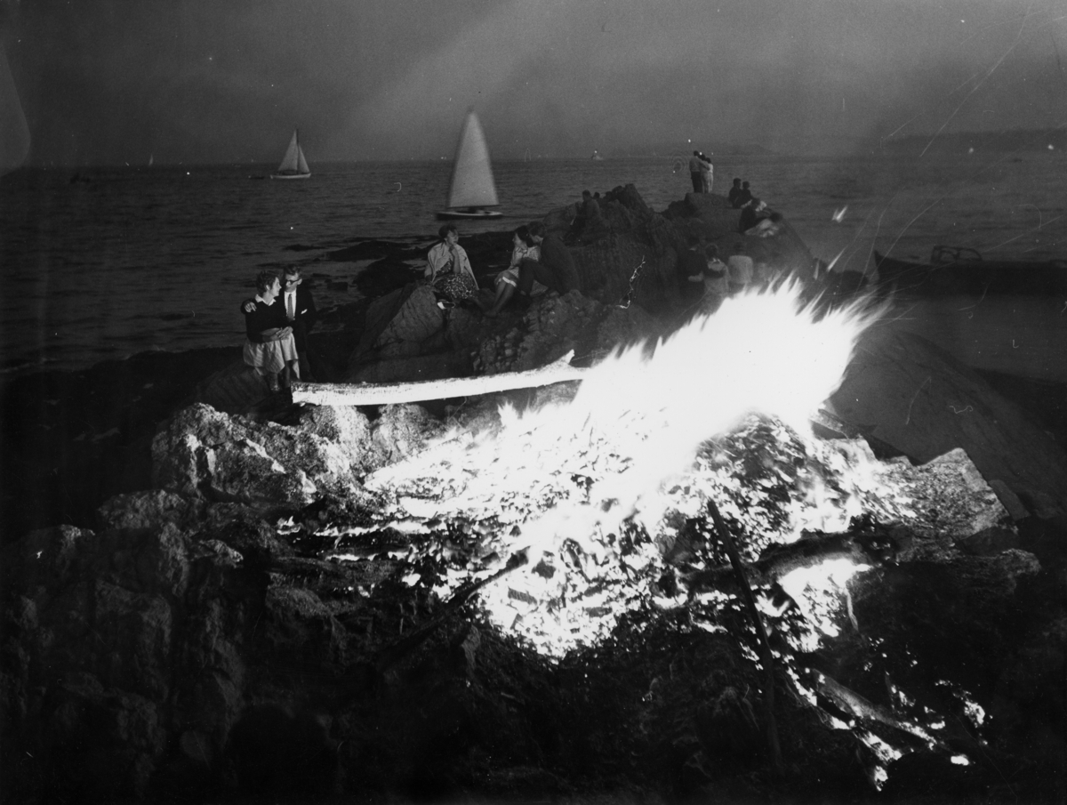 St. Hansbål på Huk. Små grupper med kvinner og menn sitter rundt bålet. I bakgrunnen sees flere seilbåter.