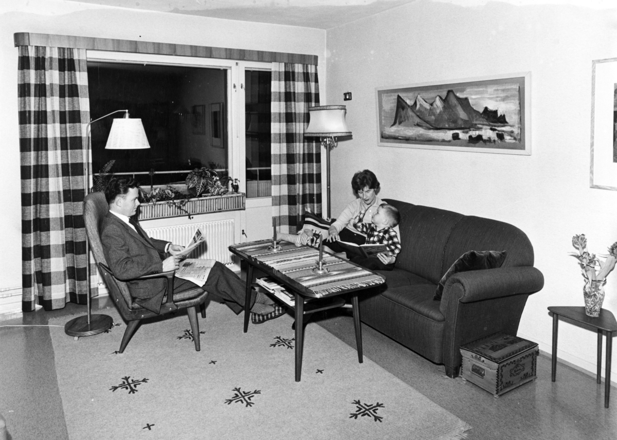 Foto av en familie som sitter i ei stue. Far leser avisa mens mor leser for gutten.