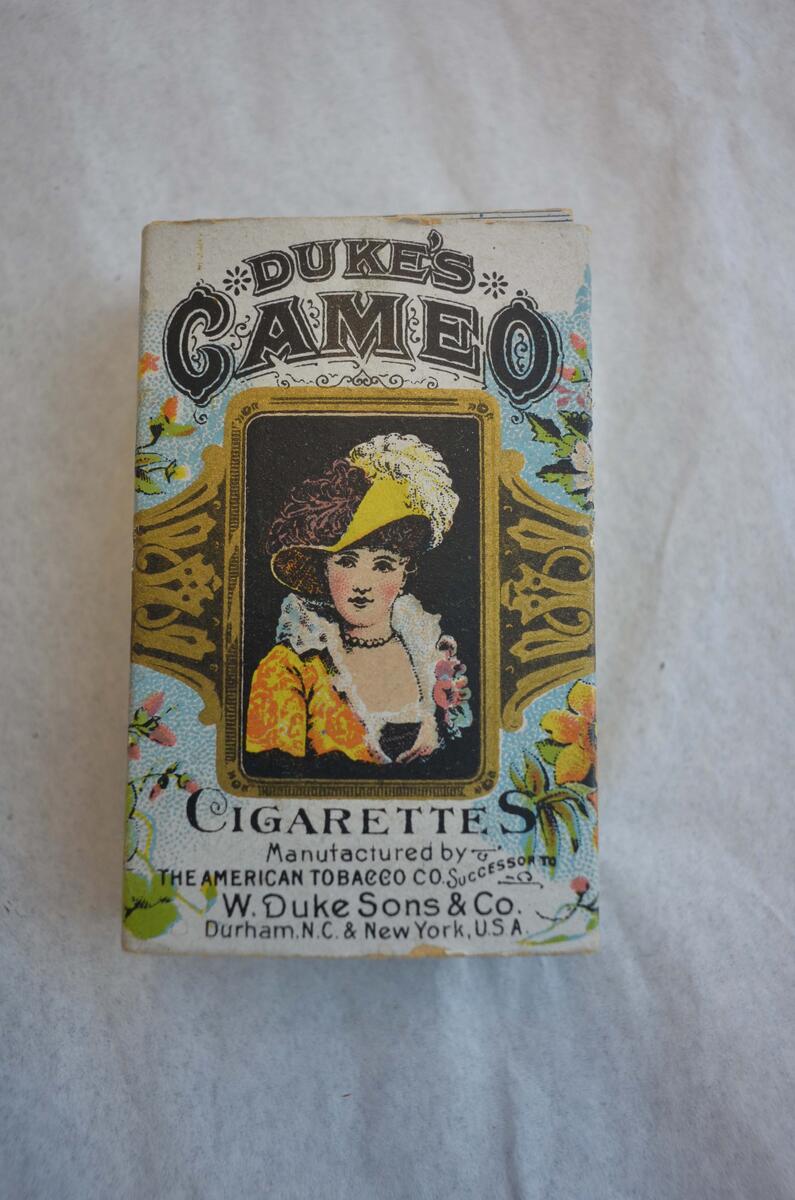 Cigarettpaket, innehållande 12 cigaretter av märket "Duke´s Cameo". Rektangulär vit ask med tryck i svart, guld och gult m.m. På framsidan en kvinnobild inom ram, ovanför vilken står: Duke´s Cameo och under bilden: Cigarettes Manufactured by The American Tobacco Co. Successor To W. Duke sons & Co. Durham. N.C. & New york, U.S.A. På sidorna och baksidan finns bl.a. målningar av blommor.