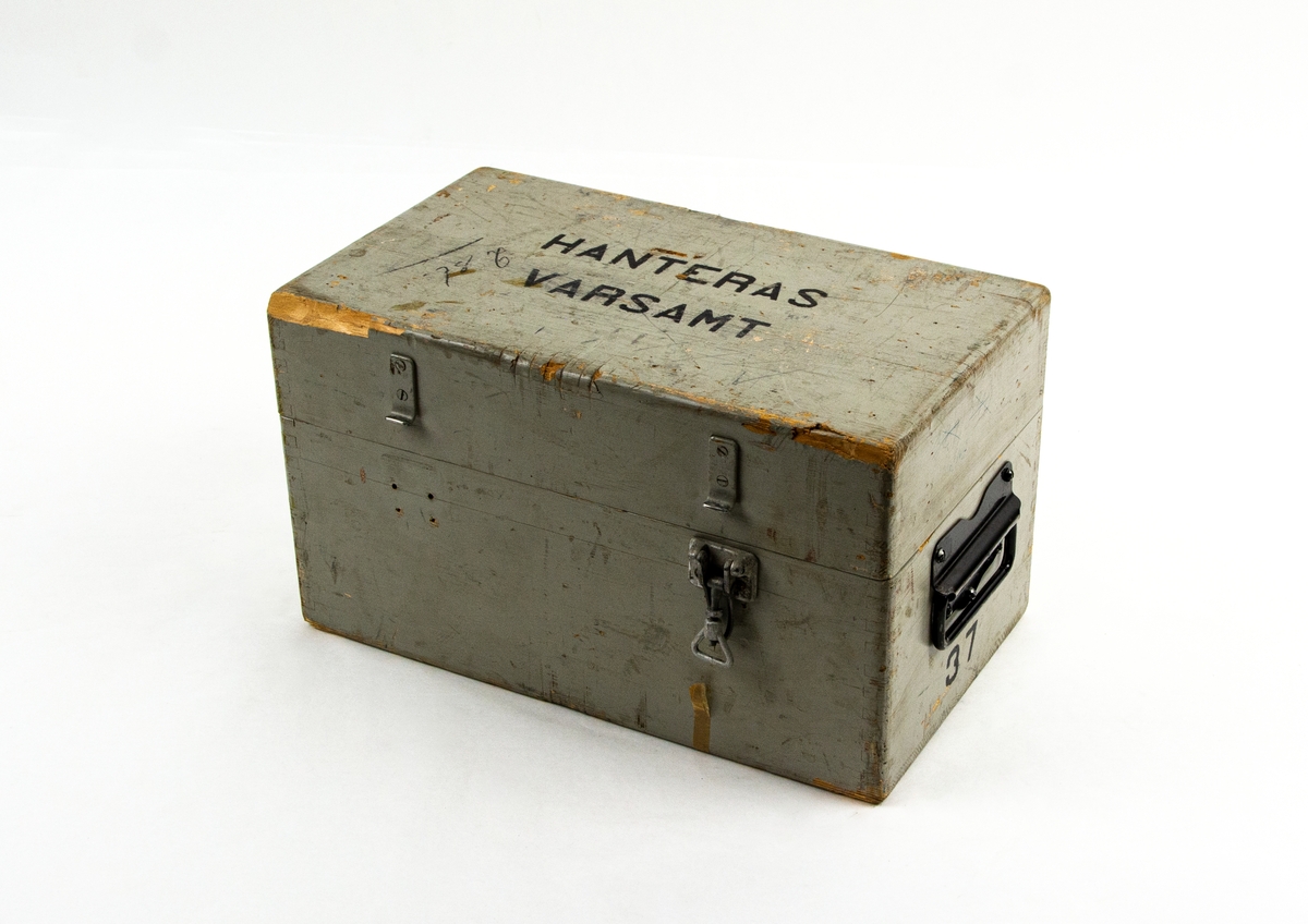 Fällmekanism BB3-1. Rektangulär box i metall. Förvaras i tillhörande transportlåda i trä. Emballaget tillhör Svenska Aeroplan Aktiebolaget Jönköping, låda nr 37.