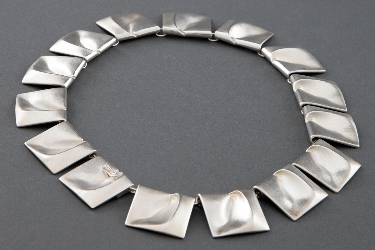 Halskjede bestående av sammenhektede kvadratiske elementer i støpt sølv. Hvert element har en abstrakt skulpturell utforming og preges av en myk organisk bevegelse.