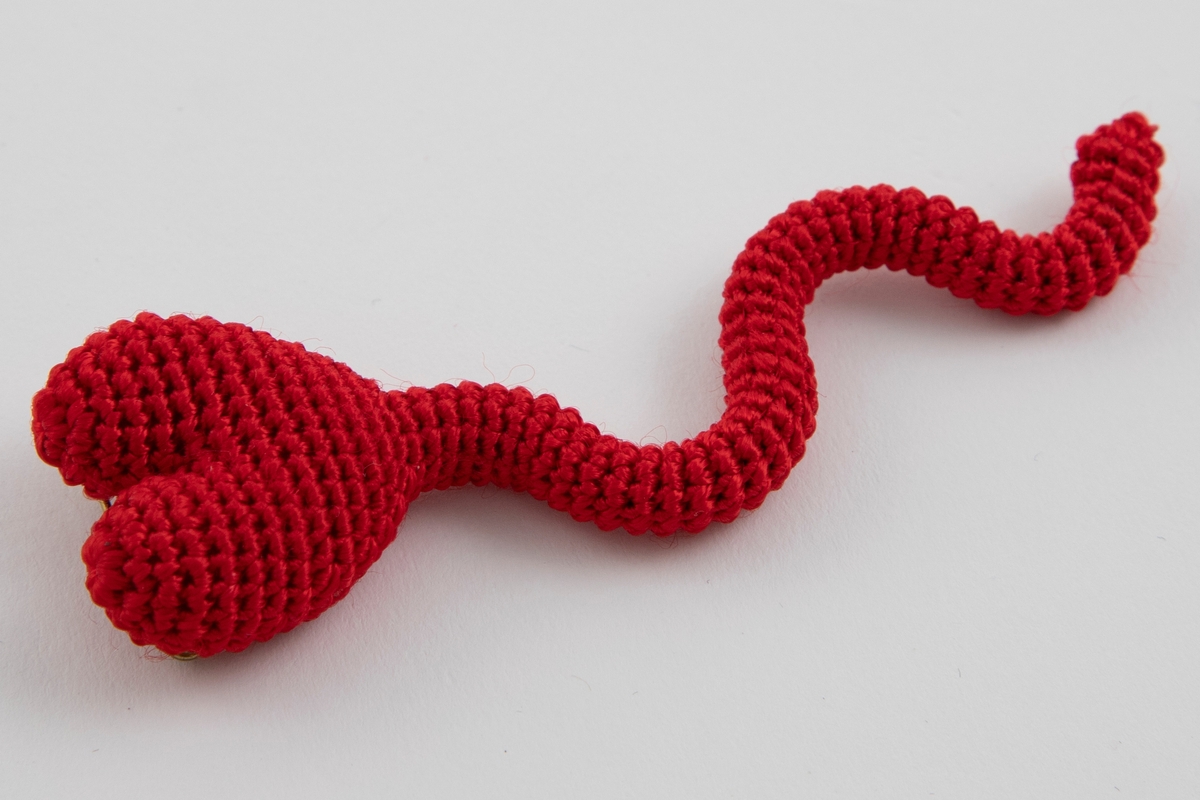 Heklet brosje i rødt garn med en bred, hjerteformet øvre del og en smal, slyngete nedre del som ligner en hale. Sikkerthetsnål på baksiden til å feste smykket.