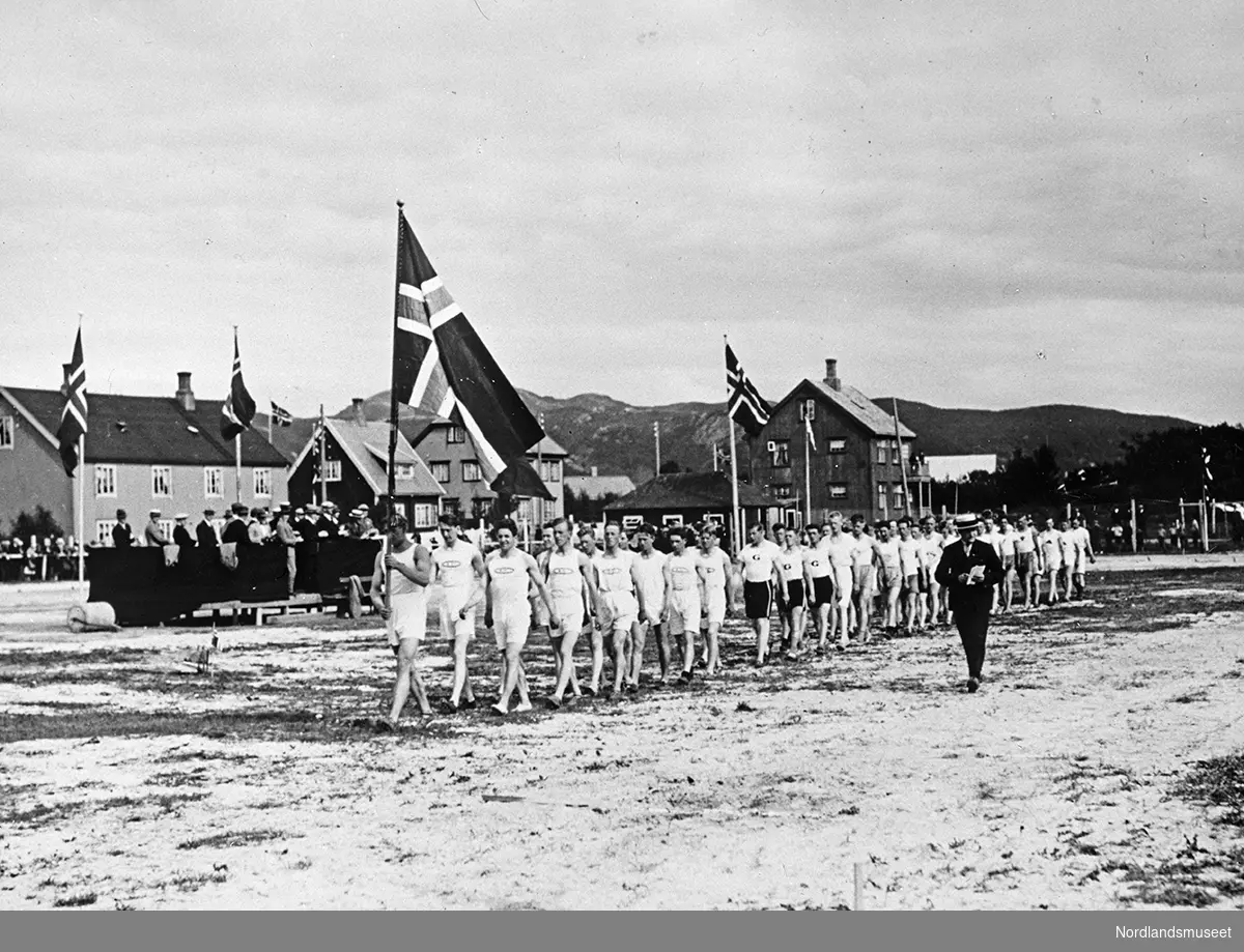 Turnere marsjerer på idrettsplassen ved sykehuset/middelskolegården (?). Bærer det norske flagg. Bygninger til venstre og flere tilskuere.