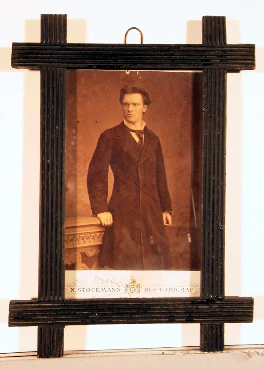 Portrettfotografi av ung mann i lang mørk jakke med hvit skjorte med opprettstående flip og løstbundet slips.
