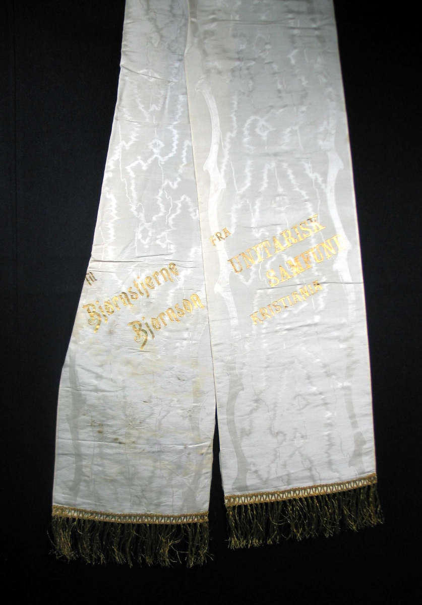 Begravelsessløyfe i hvit silke med tekst og frynser i gull.