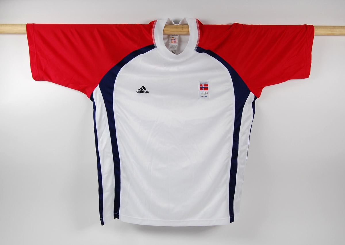 T-skjorte med hovedfargene hvit, rød og blå. Logo for adidas og påsydd merke med logo for Olympiatoppen på fremsiden. De olympiske ringene laminert på baksiden.