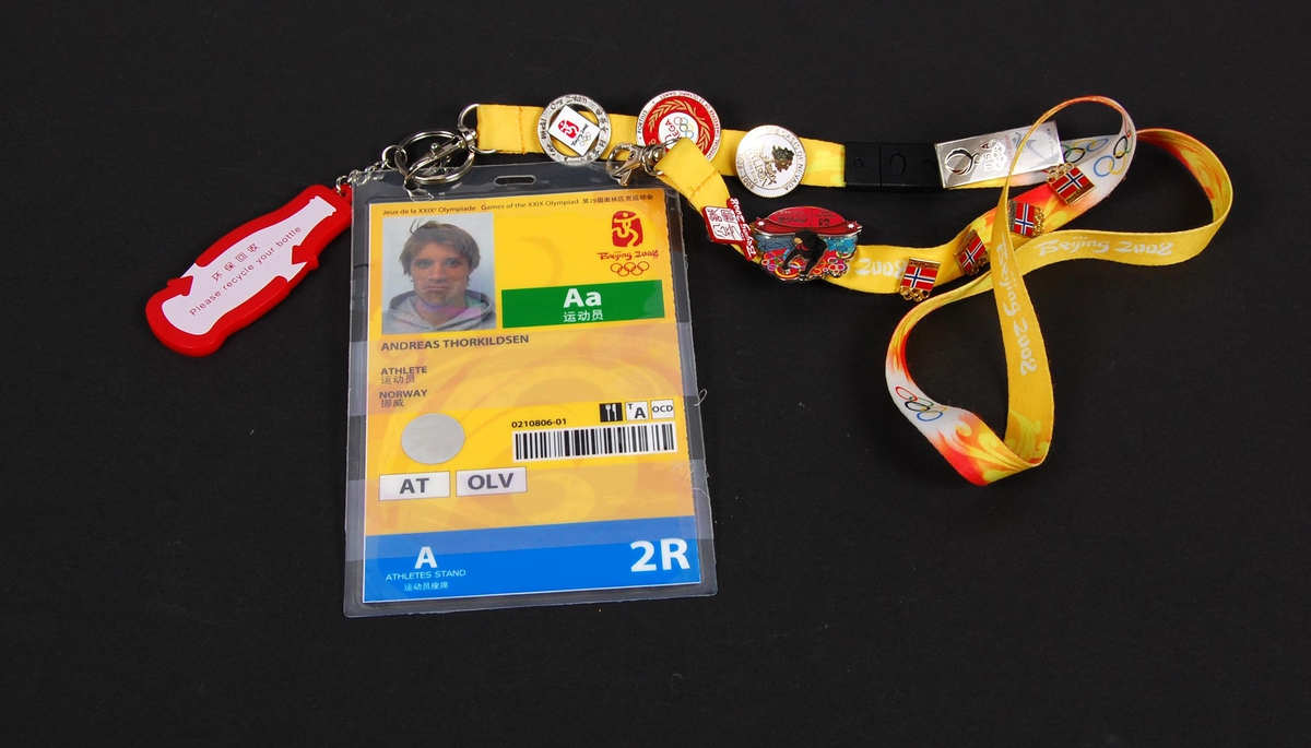 Flerfarget identitetskort med passfoto av Andreas Thorkildsen og logo for de olympiske sommerleker i Beijing i 2008. Til kortet er det festet et bånd med diverse pins laget i forbindelse med OL i Beijing.