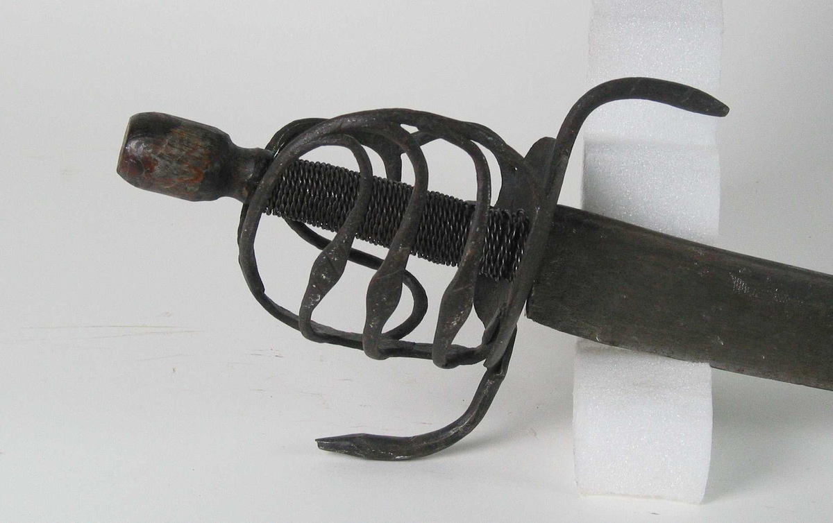 Rund tønneformet knapp i tre. Asymmetrisk feste med tre sidebøyler som har fortykninger på midten på utsiden og tommelbøyle og to sidebøyler på innsiden. Nyreformede parérplater. S-formede runde parérstenger med fortykkede avslutninger på endene. Fremre parérstang er nyere.
Tregrep med jerntrådvikling.
Enegget krum kline med smal ryggstillt blodrand. Smedmerke på venstre side. Klingespissen er dekorert med tre små hull.