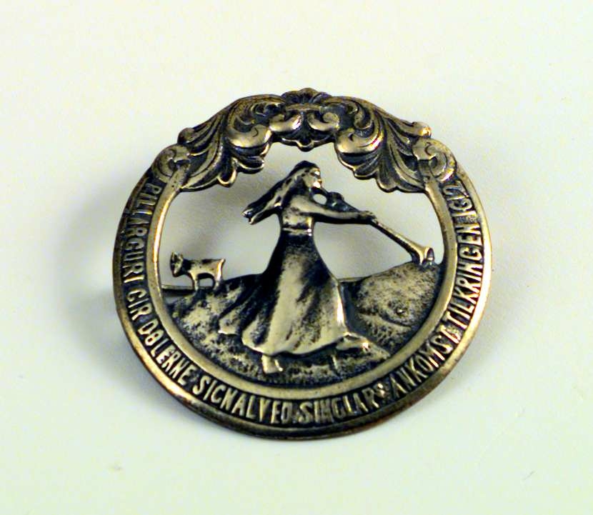 Brystnål eller brosje i sølv med Pillarguri-motiv. Nålen ligger i en eske fra gullsmed J. Frisenberg. Esken er lys blå.