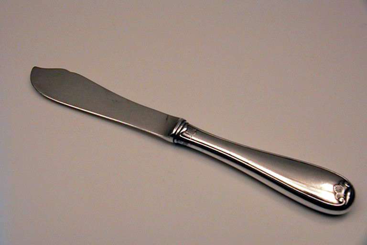 Kniv til fiskebestikk. Materialet er 90 gram sølv. Kniven har et enkelt bladmønster (viftemønster). Stempelet består av et tårn og bokstavene N (i kvadrat) og NM 90.