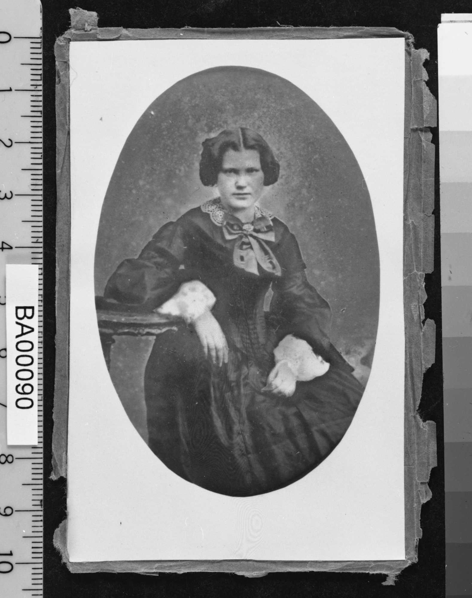 Portrettfotografi av en ung dame sittende med høyre arm hvilende på pidestalbord. Hun er ikledd en mørk kjole med snøreliv (korsett), store hvite kniplingsmansjetter og en stor sløyfe i halsen. Frisyren er oppsatt med midtskill.