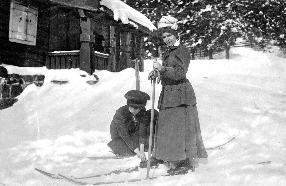 Kvinne som hjelper en kvinne med skiene, hytte i bakgrunnen, vinter