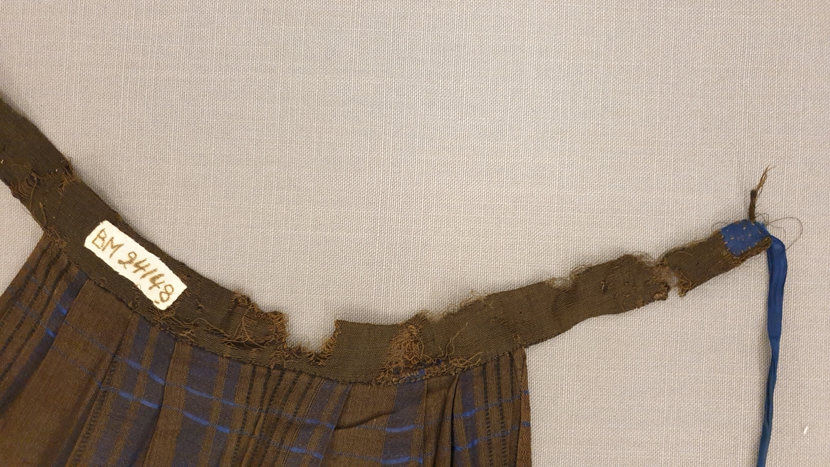 Förkläde med mörkare brun/svart varp (ull? bomull?) och satinränder i blått silke. Rynkat i midjan mot linning, knytband på höger sida. Linningen mycket trasig, vänster knytband saknas.