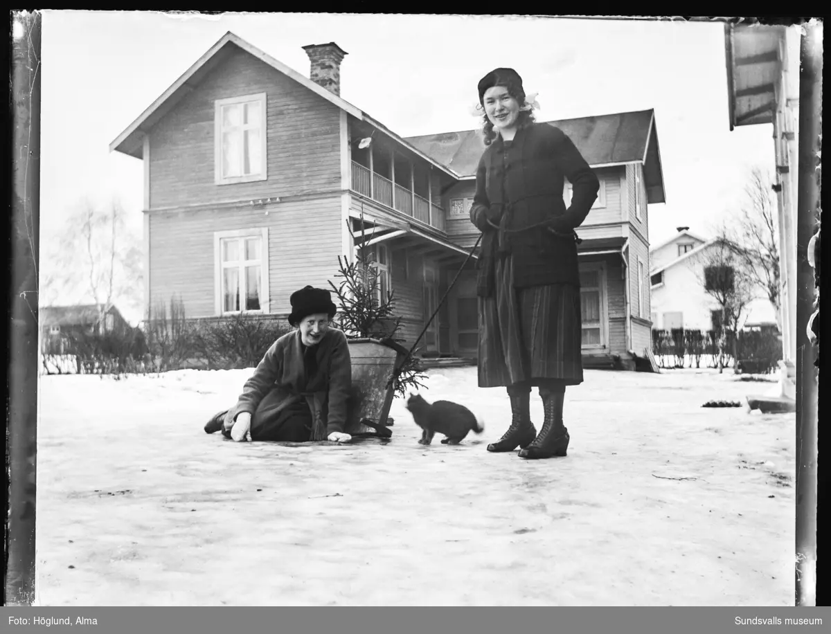 Göta Bäckström med en annan flicka och en katt på gårdsplanen till hennes hem i Fanbyn, i bakgrunden gårdshuset.