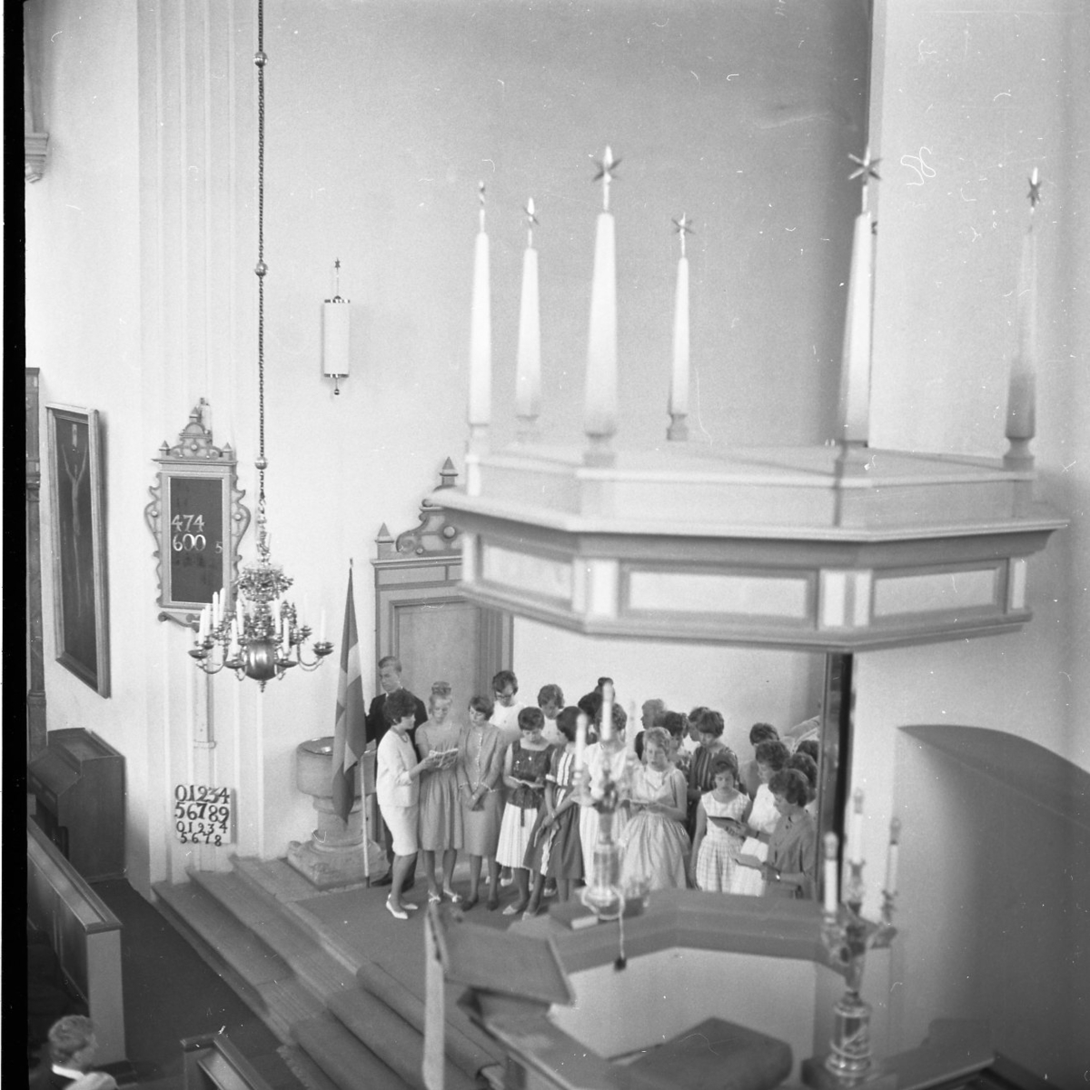 En flickkör står framför altaret i Grännakyrka och sjunger. Bredvid står en fanbärare med svenska flaggan. De skyms lite av predikstolen som finns i förgrunden.
