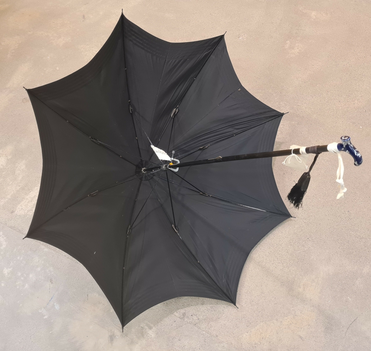 Parasoll i svart tyg. Vid skaftet hänger två tofsar.

Ingår i en samling av nio olika parasoller med olika utseende och modell.