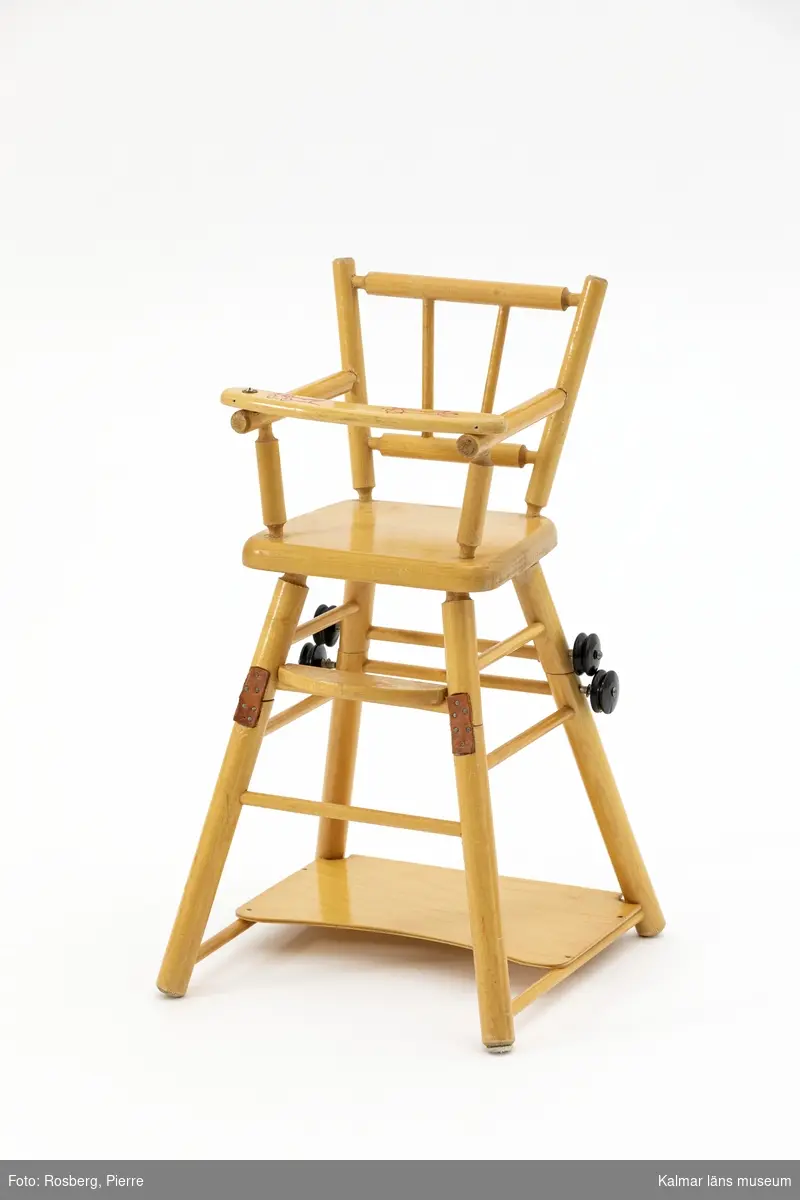 KLM 46610. Dockstol. Av trä med detaljer av läder och metall. Hög stol som går att fälla till lägre stol med bord, försedd med svartmålade hjul. På bygeln tryckt motiv med en katt och en råtta. På bygeln har det tidigare suttit en kulram.