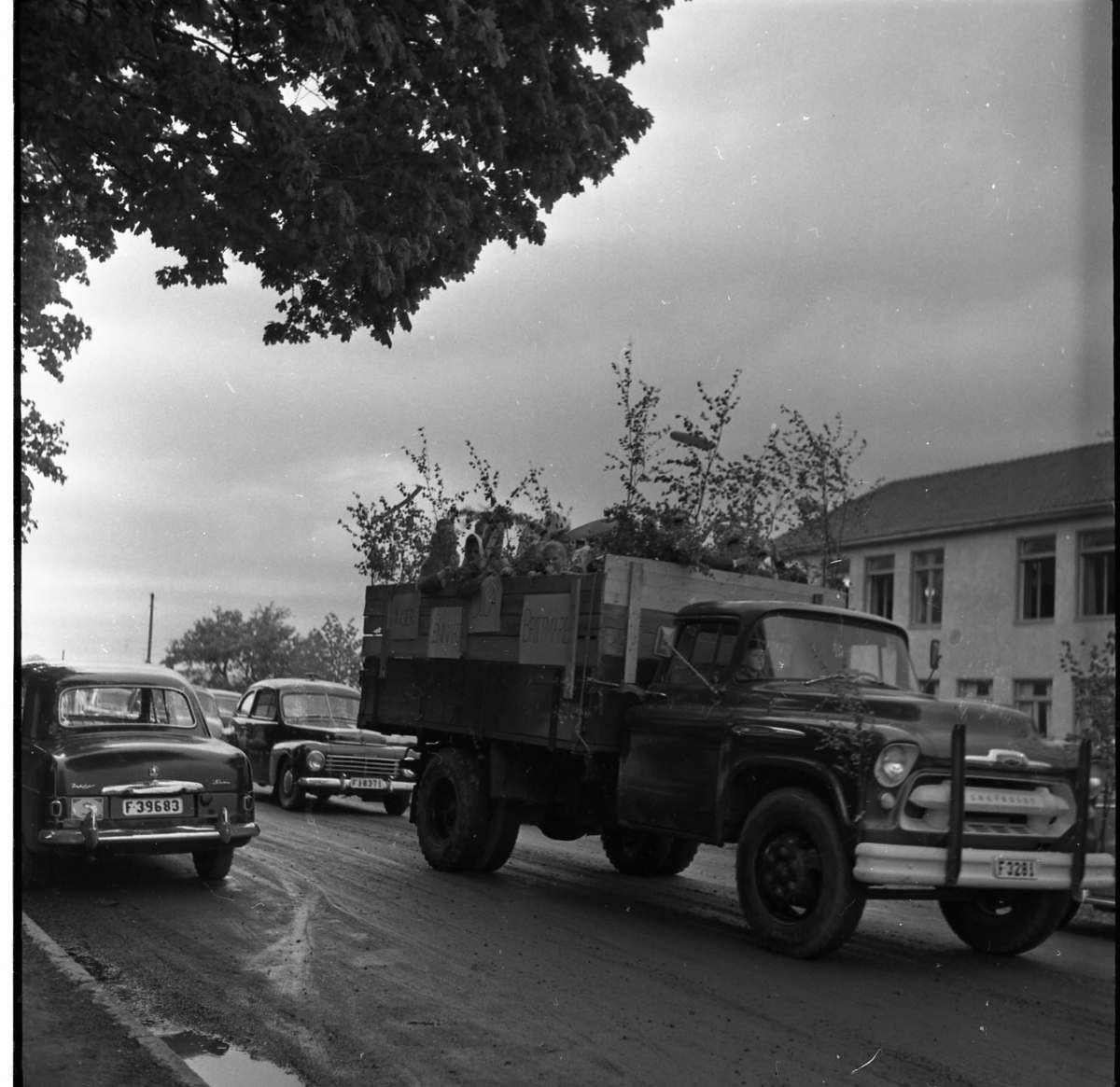 Realexamen 1961. En lastbil kör längst med en lerig gata. Lastbilen är klädd med björkris. Studenternas namn står på plakat som hänger längst med lastbilsflakets sida. På flaket står barn och studenter.
