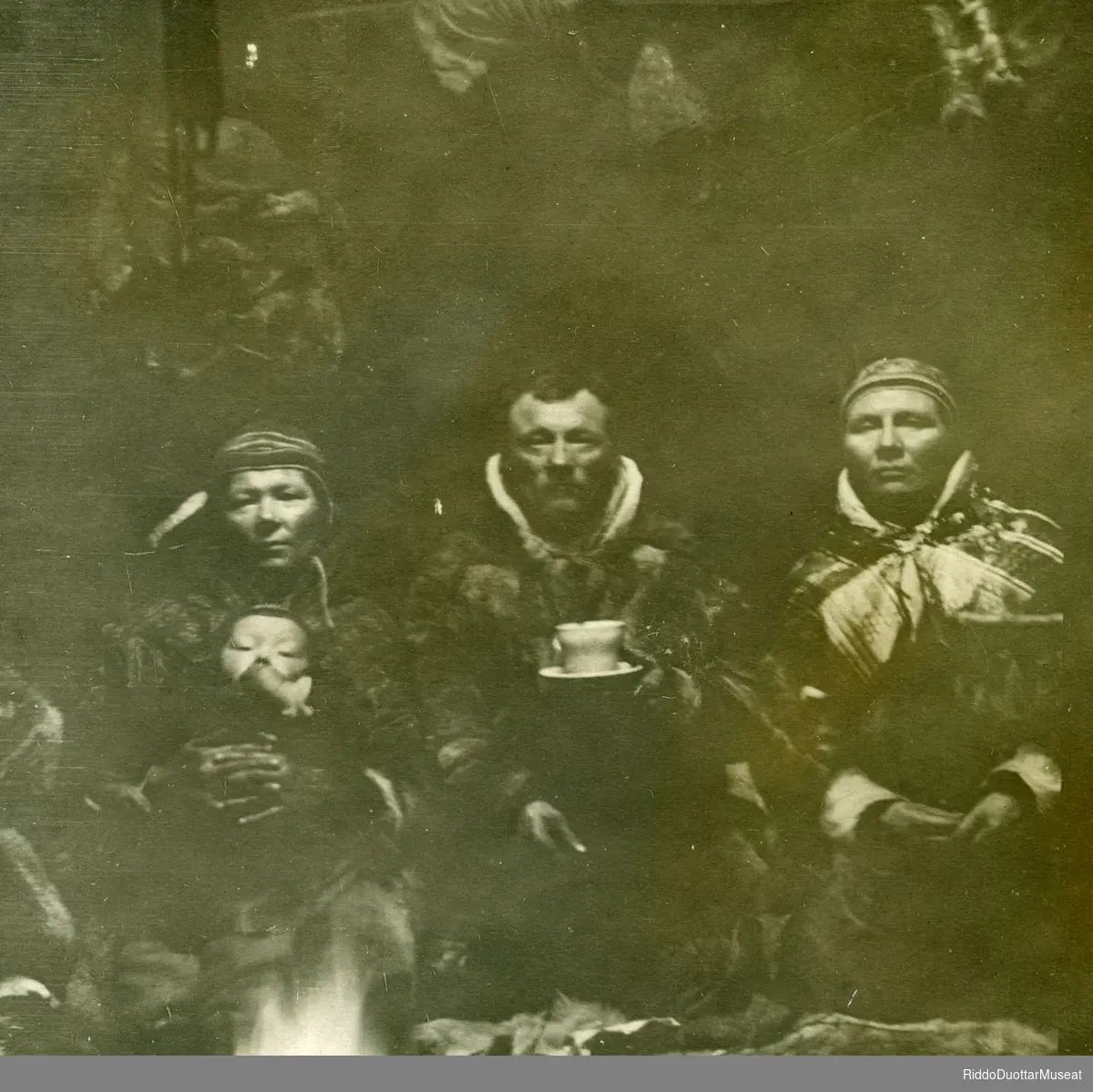 Olbmot goadis. 
Tre personer og en unge sitter ved bålet inni lavvo'en.