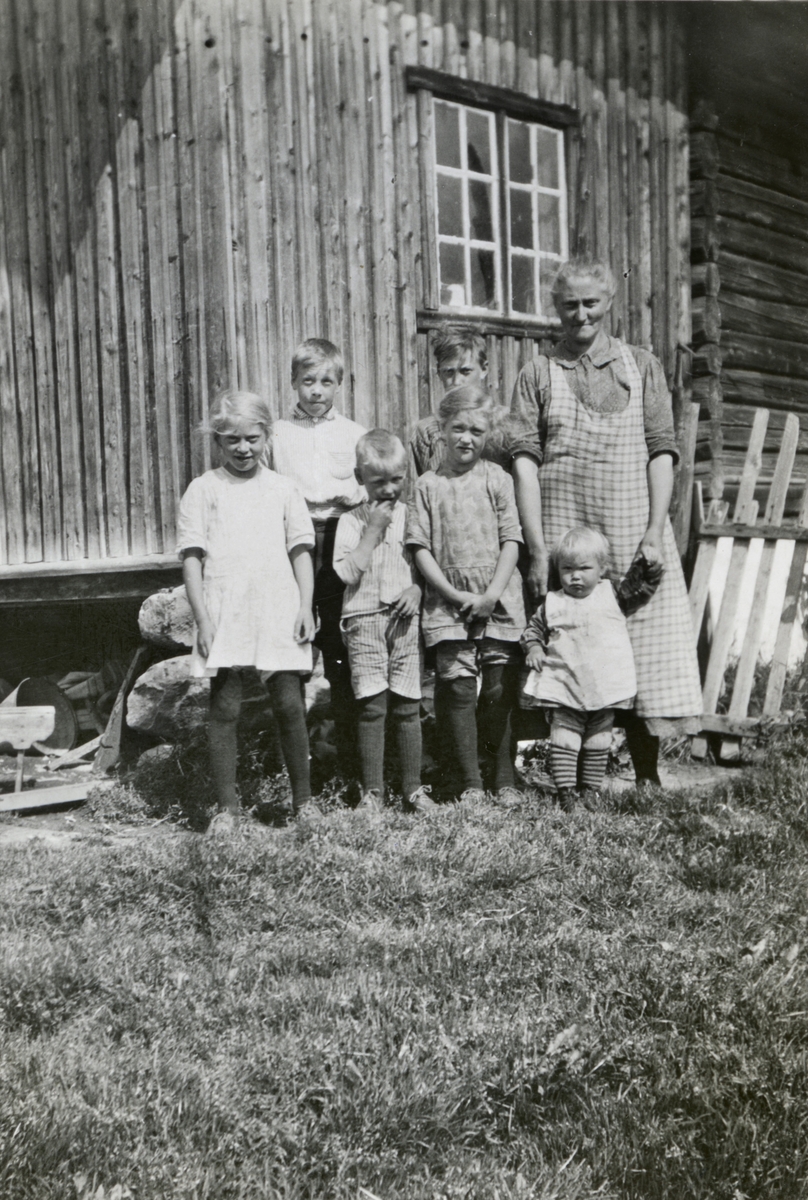 Dame og flere barn står ved tømmerhus med stakitt.