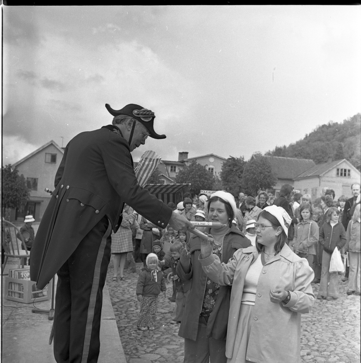 En man i borgmästaruniform står på en scen och delar ut polkagrisar till flickor. Folksamling i bakgrunden.