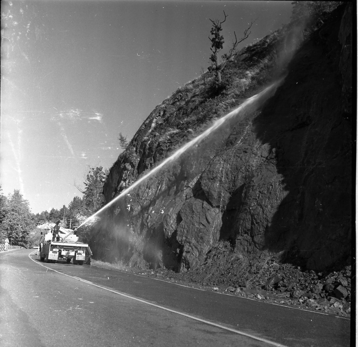 Rasberget vid länsgränsen 1961. En vattenkanon åker längst med en motorväg och har kanonen riktad mot en bergvägg. Vattnet träffar högt upp på berget och nedanför ligger rasmassor.