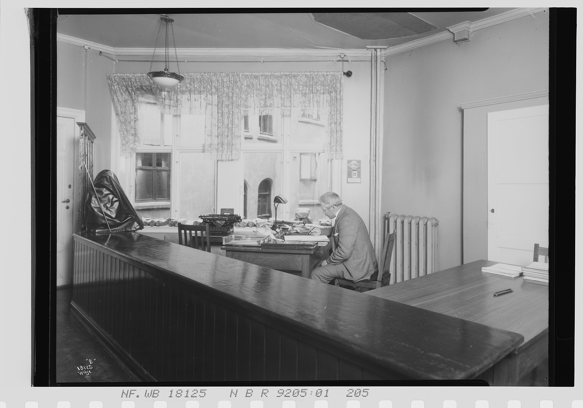 Mann sitter ved kontopult bak en disk, Vinmonopolet. Fotografert 1924.