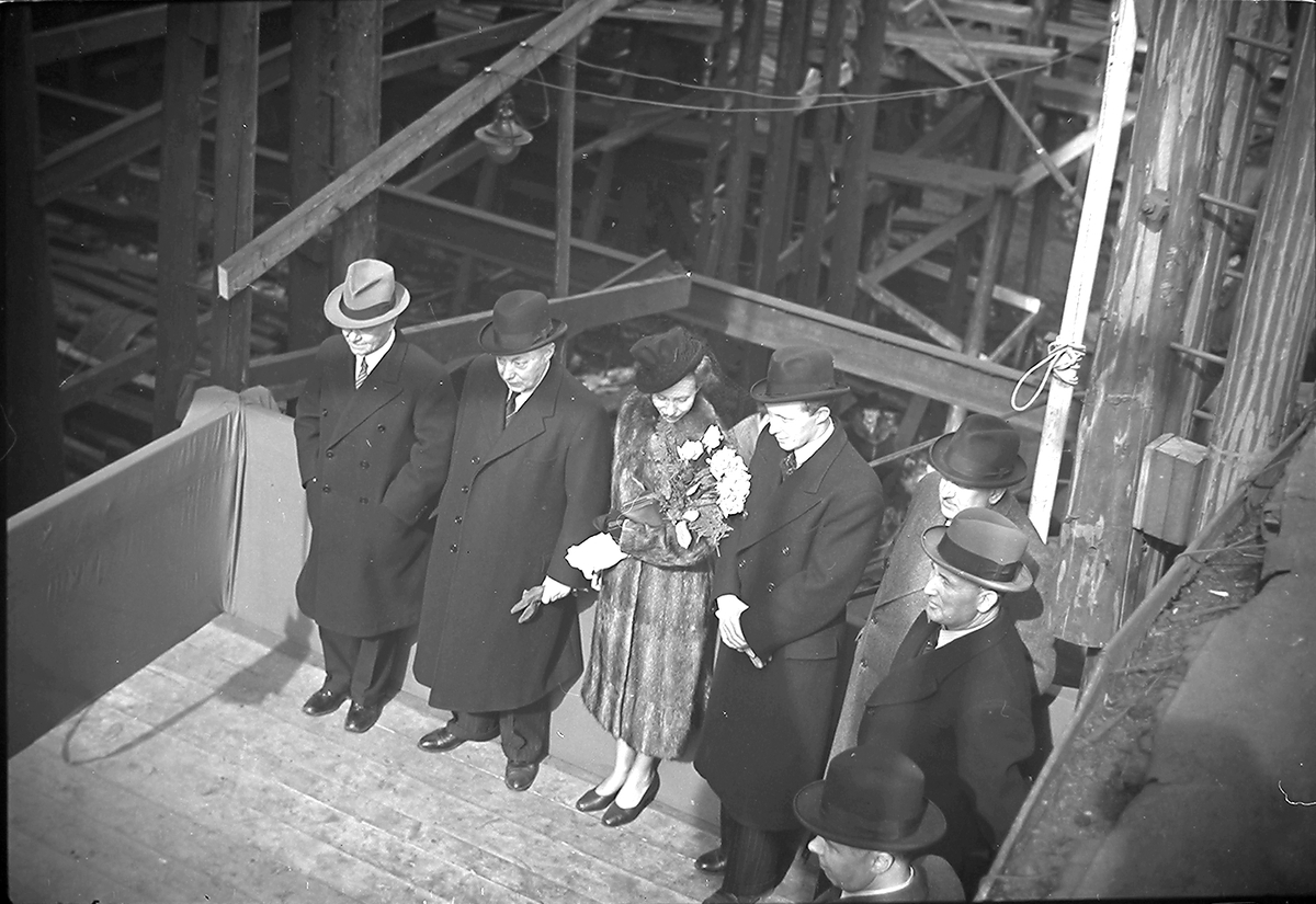 Kvinne med blomsterbukett og flere menn står i påvente av sjøsetting av et nytt skip. Fotografert 01.03.1940.