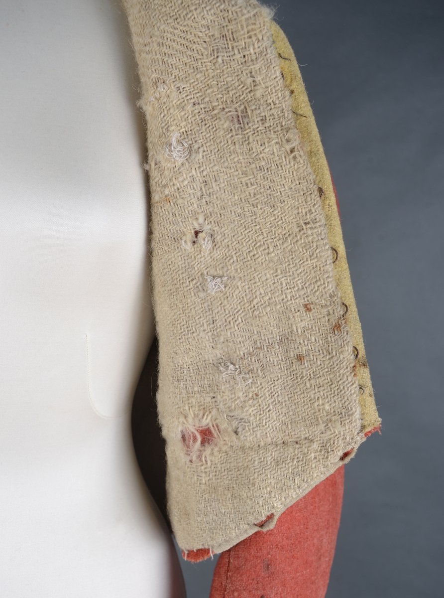 Kort trøye med høy ståkrage, sydd i grovt rødt lett valket ullstoff i empiresnitt. Trøya er helforet med grovt ullstoff. Kragen har mellomfor som er sydd fast til innerkragen. Ytterstoffet på kragen er brettet over på innsiden i øvre kant. Tittekant med rødt stoff i kortsidene på kragen. Trøya har søm MB og bakovertrekte skulder- og sidesømmer. Sidesømmene buer fra ermrigningen og mot MB. Sidesømmene slutter som legg med innlegg av gult stoff. Samme stoff brukes også som ytterkrage og slag på framstykket. Slagene er fastsydd og går opp som trekantforer mot kragen. Kneppes fast i bolen. 7 knapphull, men det mangler mange knapper. Plagget lukkes med 8 par metallhekter MF.  Nedenfor slagene er det påsydd trekanter av gråbeige ullstoff. Fra slagene foran skrår trøya bakover. Tosømsermer med dekor frampå ermene. Påsydd gult stoff med 2 striper av lys gråhvite bånd som går opp i en spiss mot innersømmen på ermet. Splitt ved yttersømmen. Denne lukkes med trekt rød knapp og knapphull. Mangler knappen på venstre erme. Stikninger langs slagene og kragen. Trøya er helt håndsydd og ytterkantene er sydd med sårkant på både for og stoff Tekst på 1 knapp, 3 knapper med stripe effekt inn mot midten.
Rød stoffhempe i nakken MB. Trøya har trolig tilhørt militæruniform fra tidlig 1800tall.