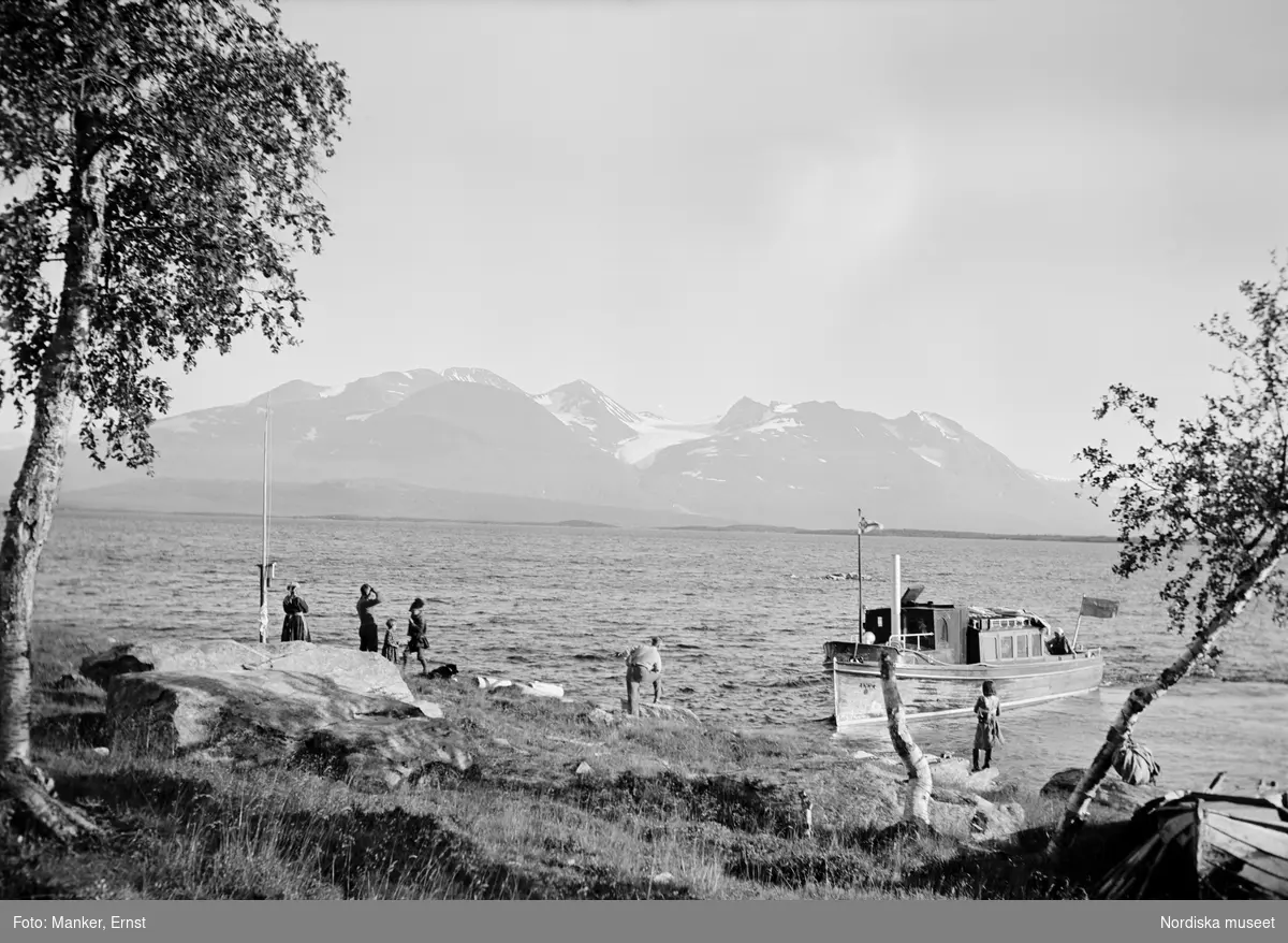 Samer och turister i samspråk vid båtlänningen vid Sörkaitumsamernas sommarviste i Ritsemjokk. Båten Akka har lagt till vid stranden, fjället Akka syns i bakgrunden.