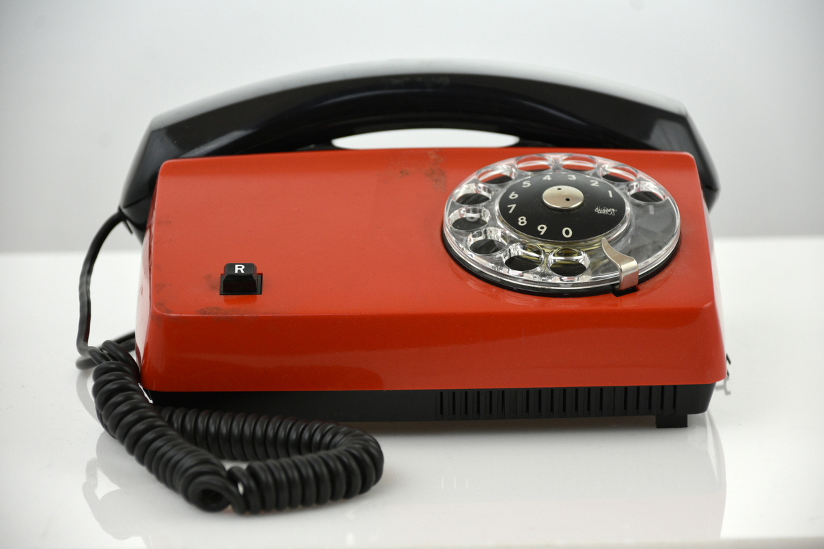 Telefonapparater Diavox 2 st, tillverkade år 1980, för AT-system, huvudapparat, anknytningsapparat till abonnentväxel och sidoapparat. Bordsmodell av plast med röd kåpa, svart mikrotelefon, fingerskiva, R-knapp för aktivering av tilläggstjänster under pågående samtal, högohmig reglerbar ringklocka, elektretmikrofon och elektronisk talkrets med automatisk dämpningskompensering.
