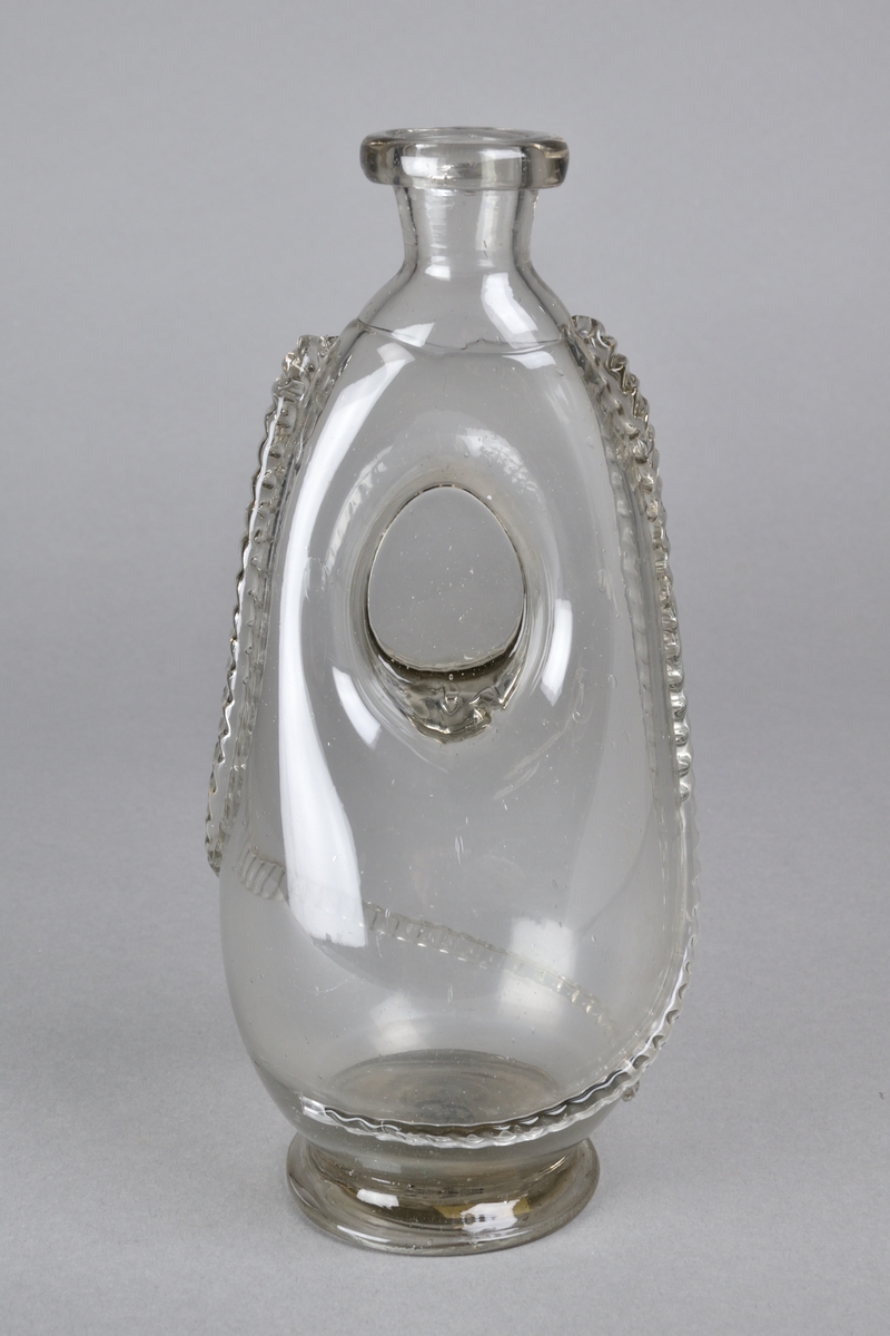 Flaska tillverkad i ofärgat glas. Bukig nedtill, med ett hoptryckt parit på mitten. Smal och kort hals, med en bred ring runt mynningen. Dekorerad med ett påsatt, räfflat glasband. Flaskan står på en rund fot. 